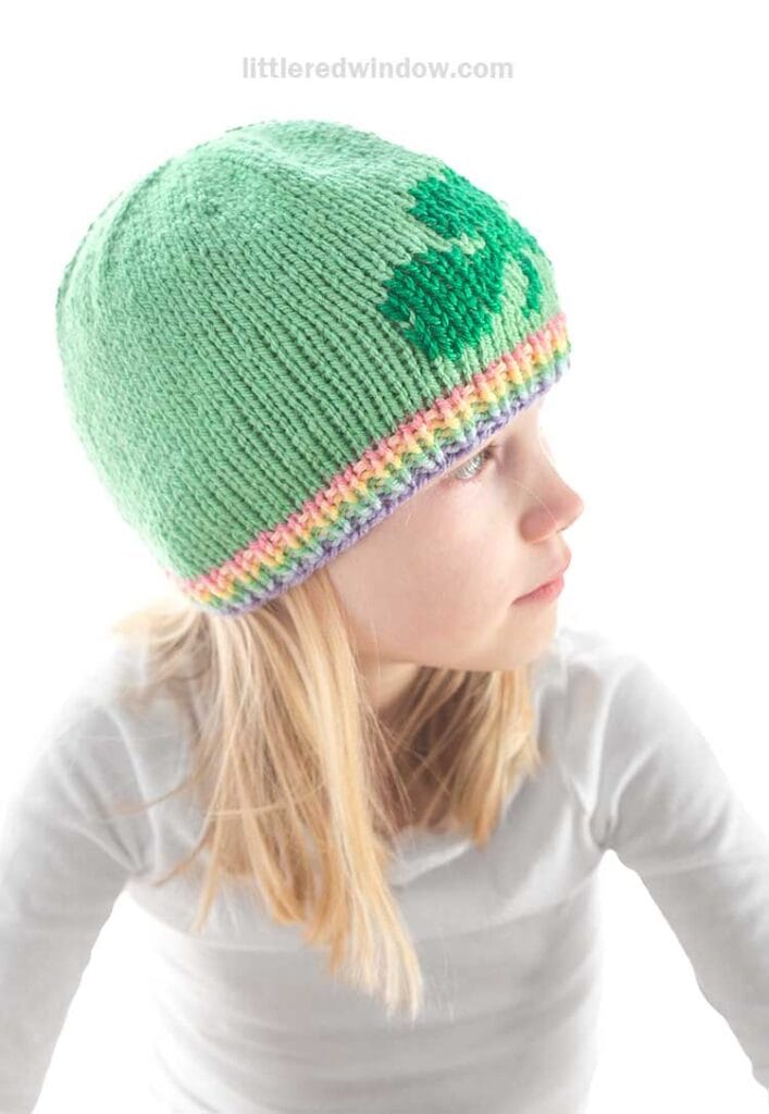 A child is sporting a knitted green shamrock hat, featuring a darker heart pattern and a multicolored rainbow brim. The child is facing sideways in a light gray shirt against the bright white background.