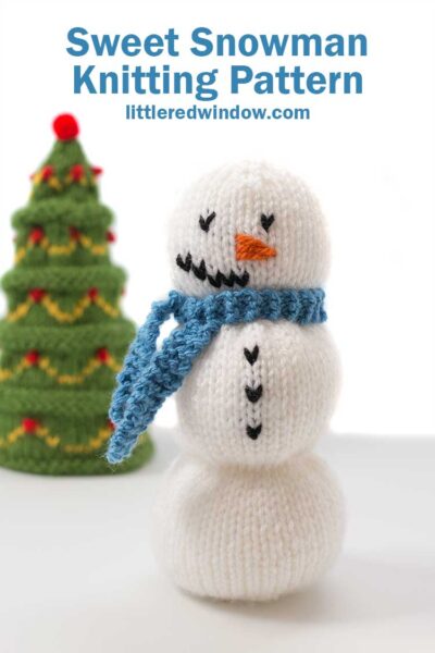 white knit snowman with a blue knit scarf standing on a white background in front of a green knit Christmas tree