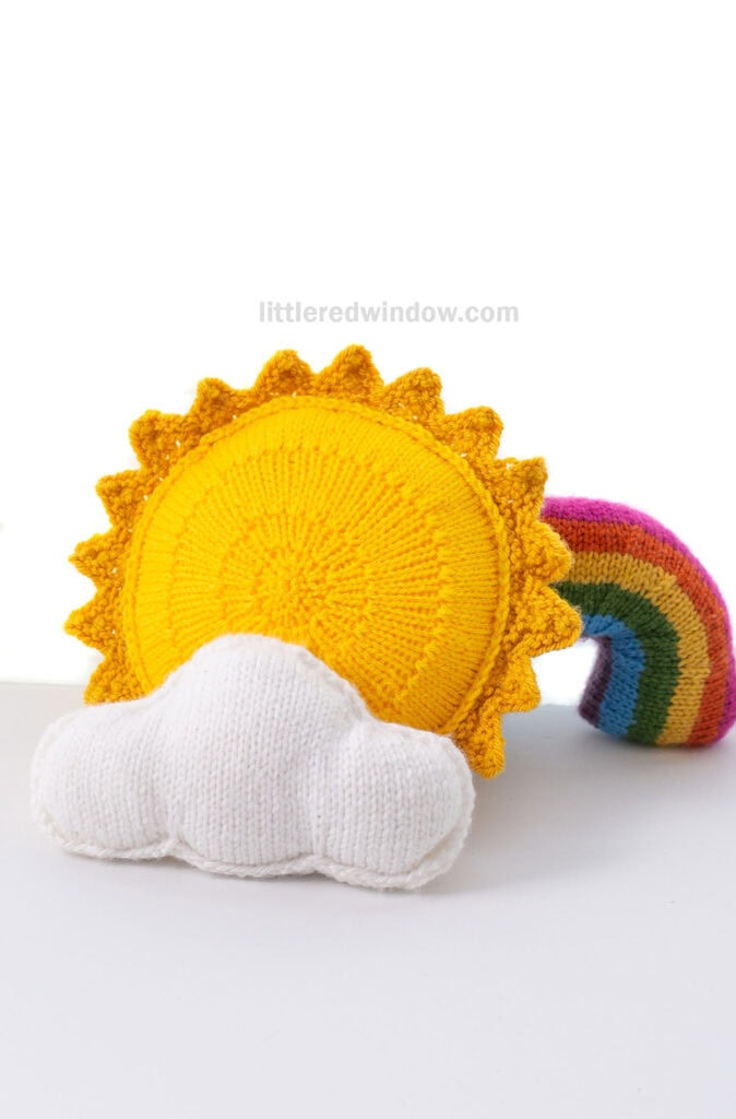 Bright yellow knit sun with a knit cloud and rainbow in front and behind in in front of a white background