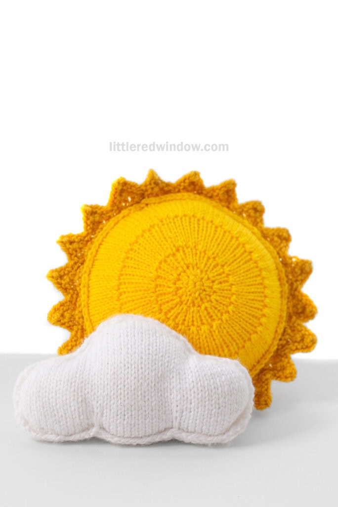 bright yellow knit stuffed sun shape with a knit white cloud shape in front of it on a light fray background