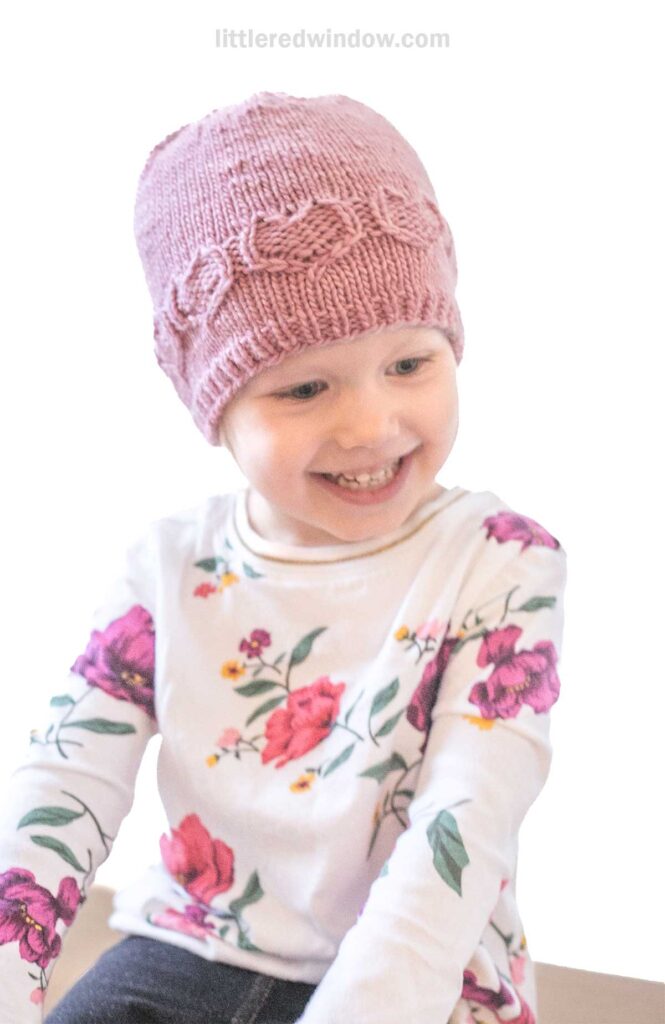 A child smiling, wearing a sweetheart pink knitted hat and a white floral shirt, against a plain background.