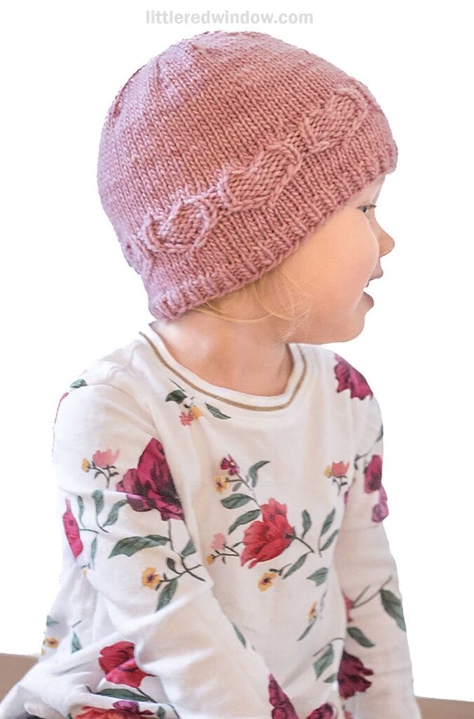 A child wearing a pink sweetheart hat with a braided pattern is seen from the side. They have on a white long-sleeve shirt adorned with red and green floral designs. The background is plain white.