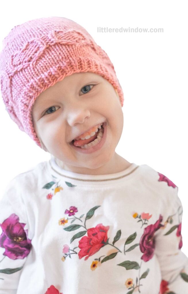 A smiling child wearing a pink sweetheart hat and a white shirt with colorful floral patterns looks playfully at the camera against a plain white background. Text