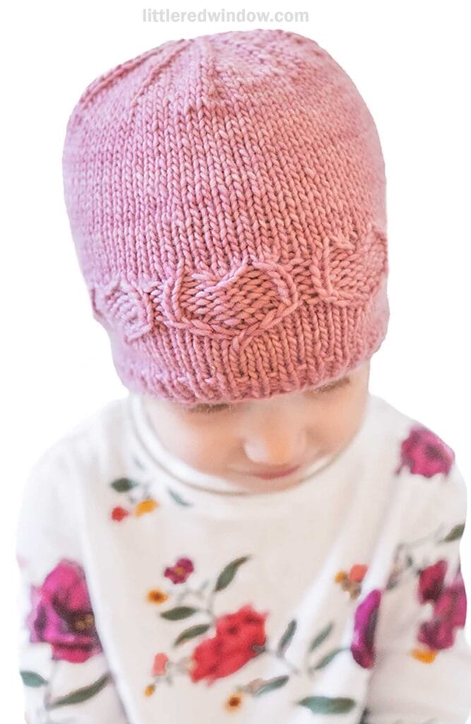 A child wearing a sweetheart hat with a textured pattern sits facing down. They are dressed in a white shirt with colorful floral prints.