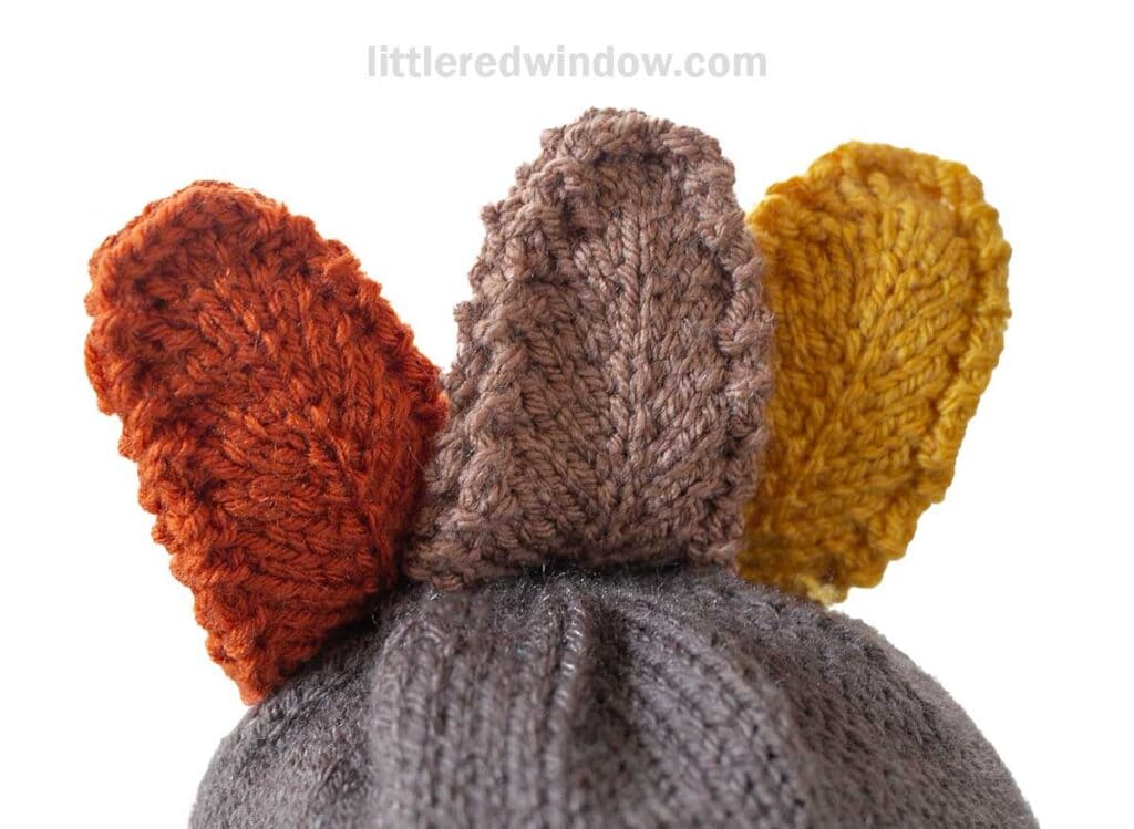 A knitted gray hat with three colorful leaf-shaped adornments on top, in shades of orange, brown, and yellow.