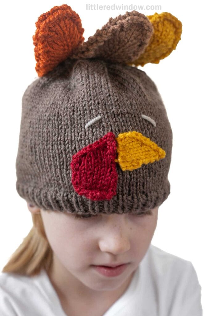 Child wearing a brown knit hat decorated to look like a turkey, with colorful knit features like eyes, a yellow beak, red wattle, and orange feathers on top. The child looks down with a neutral expression.