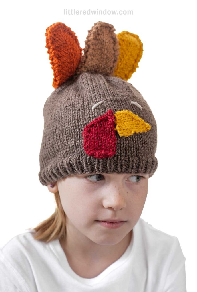 A child wearing a brown knitted hat with red, yellow, and orange accents resembling a turkey's features. The hat has patterns imitating a beak and feathers. The child is looking to the side and wearing a white shirt.
