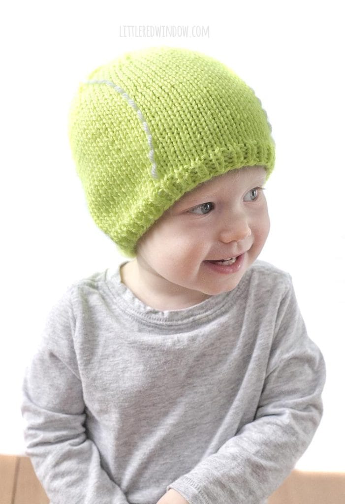 cute baby in gray shirt looking off to the right and wearing a bright green tennis ball hat