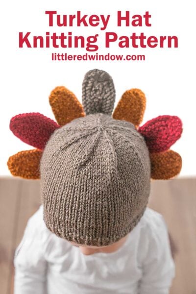 A child wearing a knit hat resembling a turkey, complete with colorful tail feathers in shades of red, orange, and brown. The text above reads "Turkey Hat Knitting Pattern" and "littleredwindow.com". The Turkey Hat is perfect for festive fall fun!