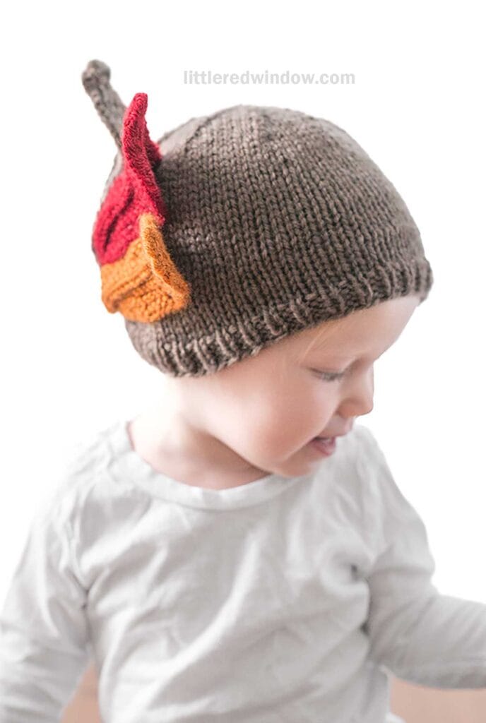 A young child wearing a brown knit Turkey Hat adorned with red and orange autumn leaves looks down and to the side. The child is dressed in a plain white shirt. The background is white, and the text
