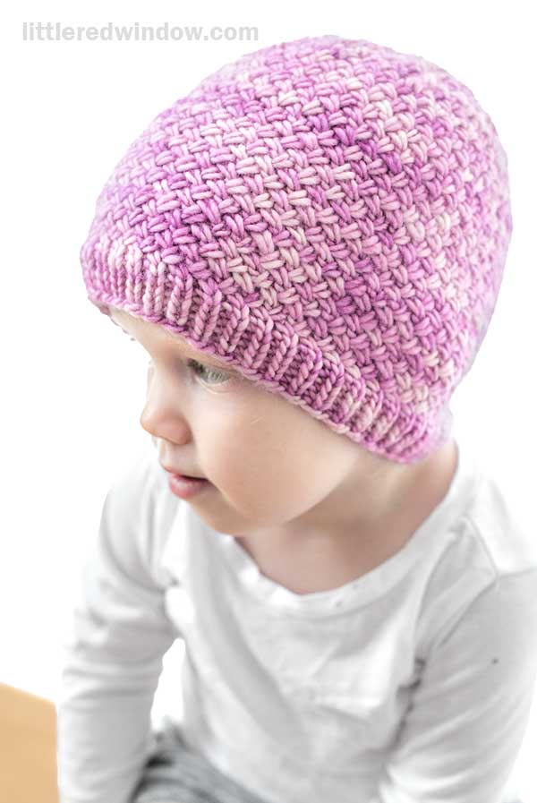 A young child wearing a hand-knitted, pink and white Tiny Basketweave Hat looks to the side. The child is dressed in a long-sleeve white shirt, set against a bright, plain background.