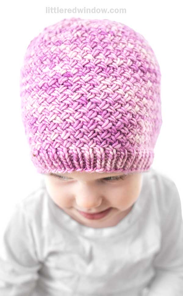 A small child wearing a light pink and purple Tiny Basketweave Hat with a textured pattern looks down and smiles slightly, set against a white background.