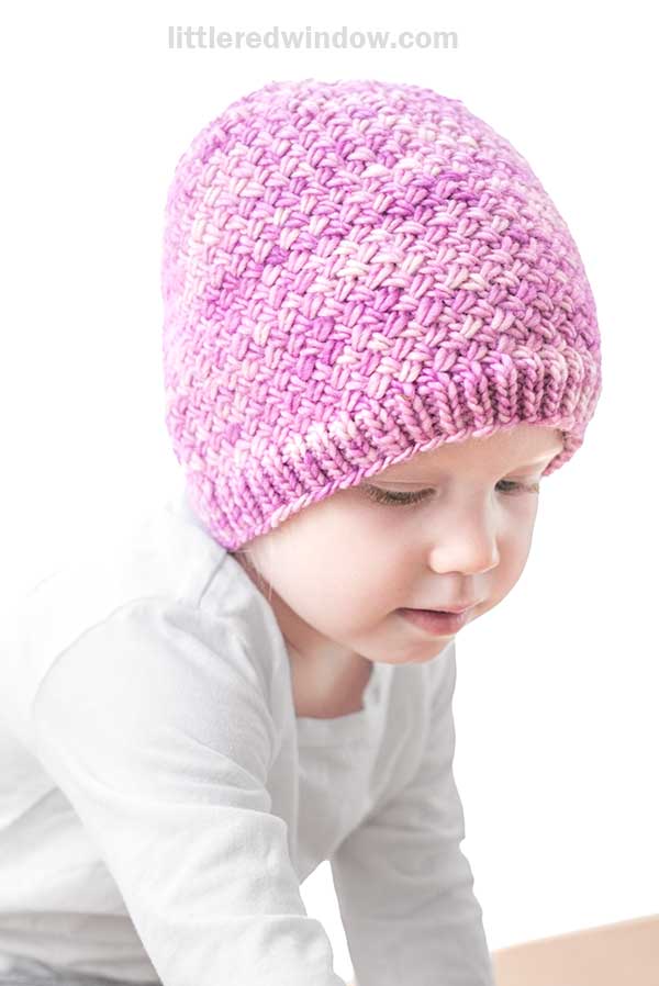 A young child wearing a pink Tiny Basketweave Hat looks down while dressed in a white long-sleeve shirt against a white background.