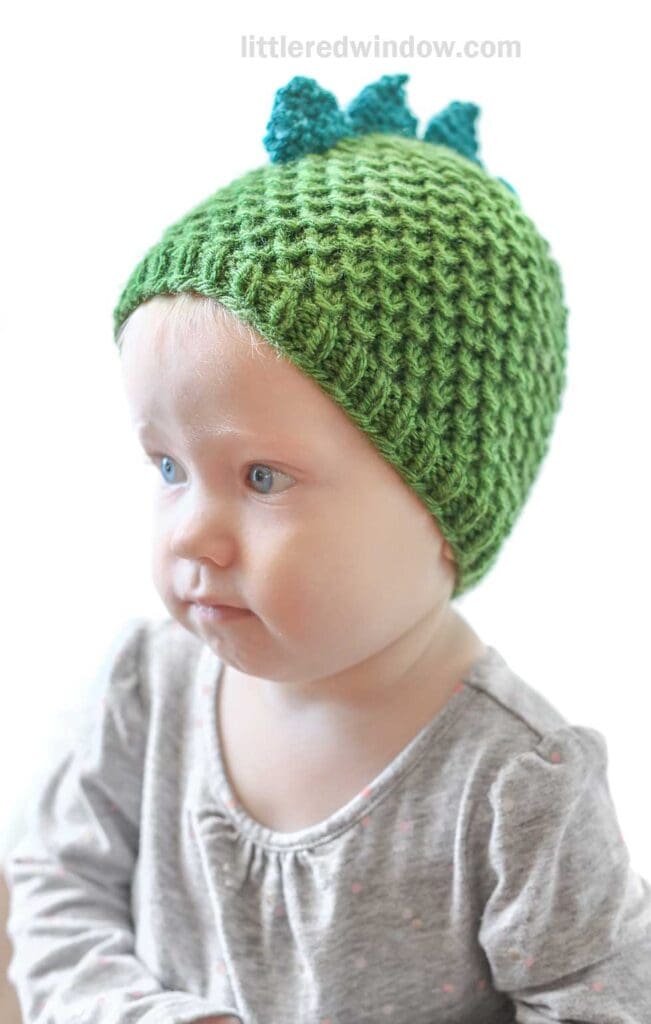 A baby wearing a green crocheted dragon hat with blue spikes looks to the side. The baby is dressed in a light-colored top with small dots. The background is plain and white, placing the focus on the baby and the hat.