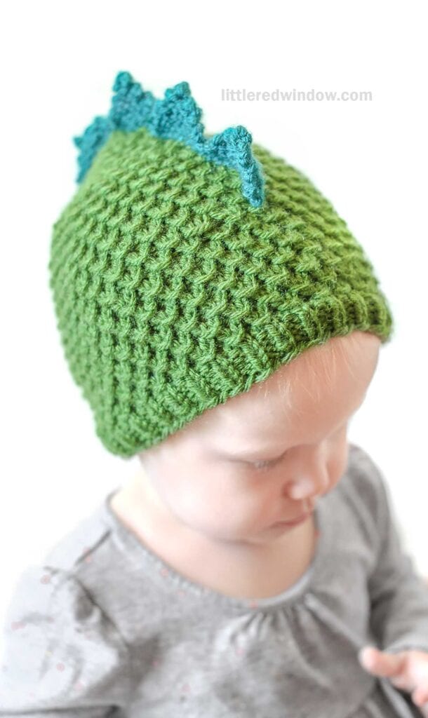 A baby is wearing a green crocheted dragon hat with blue dinosaur-like spikes on top. The baby is looking down, dressed in a light grey outfit. The background is plain white and the words