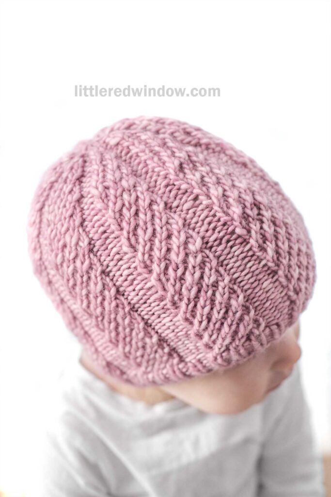 A close-up of a young child wearing a knitted pink Twisted Rib Hat with a textured pattern. The child is looking down and to the side, wearing a light-colored top. The background is blurred white, highlighting the softness of the hat’s fabric. The words