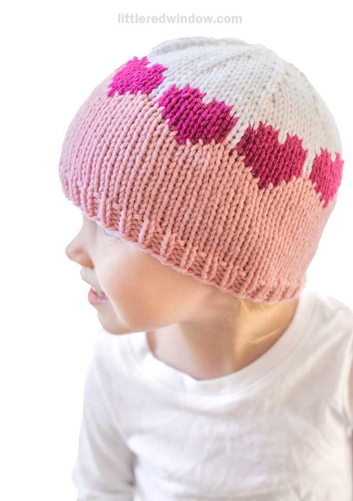 A child wearing a pink and white colorblock hat with heart patterns looks to the left. With a joyful expression, the child is dressed in a white long-sleeved shirt against a plain white background, showcasing an adorable knitting pattern that'll melt your heart.
