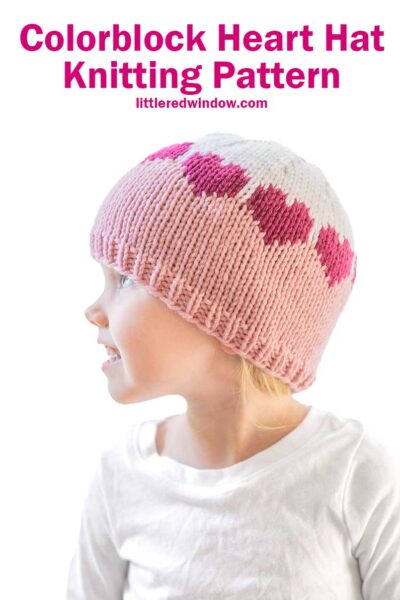 A young child dons a charming colorblock hat with pink and white hues, adorned with a lovely heart design. Glancing to the side in a crisp white shirt, they capture warmth and whimsy. Above reads, "Colorblock Hat Knitting Pattern" from littleredwindow.com.
