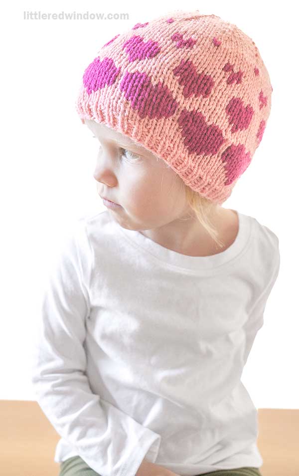 A young child, donning a lovingly hand-knit pink beanie adorned with purple hearts, sits looking to the side in a white long-sleeve shirt. The background is plain and bright.
