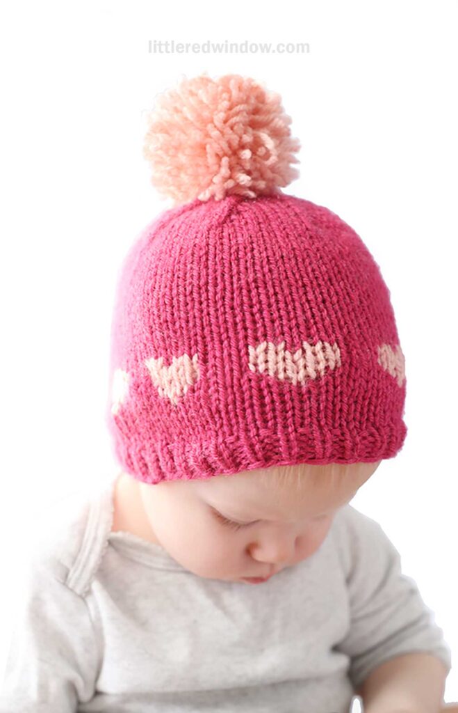 A baby wearing a heart hat with small pink hearts and a large fluffy pom-pom on top. The little one gazes downward, set against a plain white background.