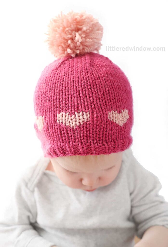 A baby wearing a charming heart hat with a knitted pink design, topped with a large pom-pom and adorned with delicate light pink hearts around the brim. Dressed in a light gray outfit, the baby gazes downward against a plain white background.