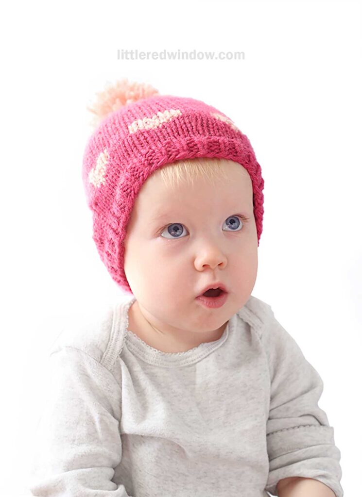 A baby, wearing a pink heart hat adorned with a pom-pom, gazes upwards. Dressed in a light gray shirt, the little one is set against a plain white background.