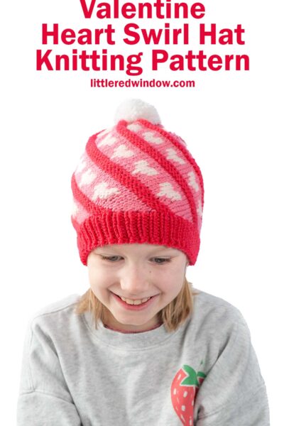 Child wearing a knitted red hat with white heart patterns and a white pom-pom on top. The child is smiling and wearing a light gray sweater with a strawberry design. Text above reads "Valentine Heart Swirl Hat Knitting Pattern"