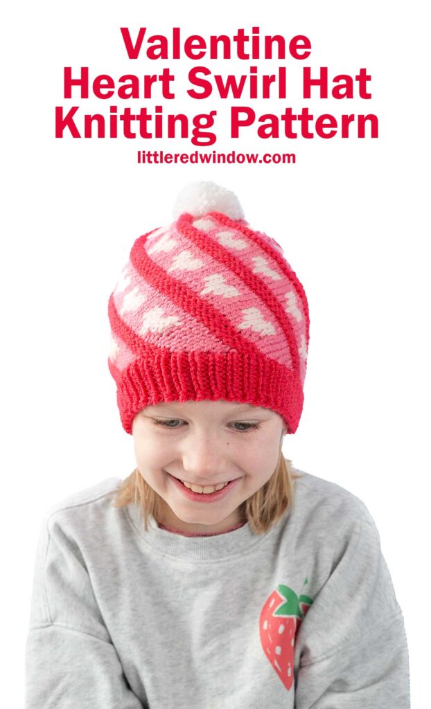 Child wearing a knitted red hat with white heart patterns and a white pom-pom on top. The child is smiling and wearing a light gray sweater with a strawberry design. Text above reads