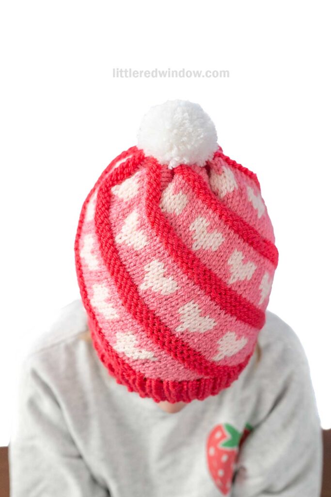 Child wearing a pink swirl knit hat with white heart patterns and a white pom-pom on top. The child wears a light gray sweater with a small strawberry graphic. The background is plain white.