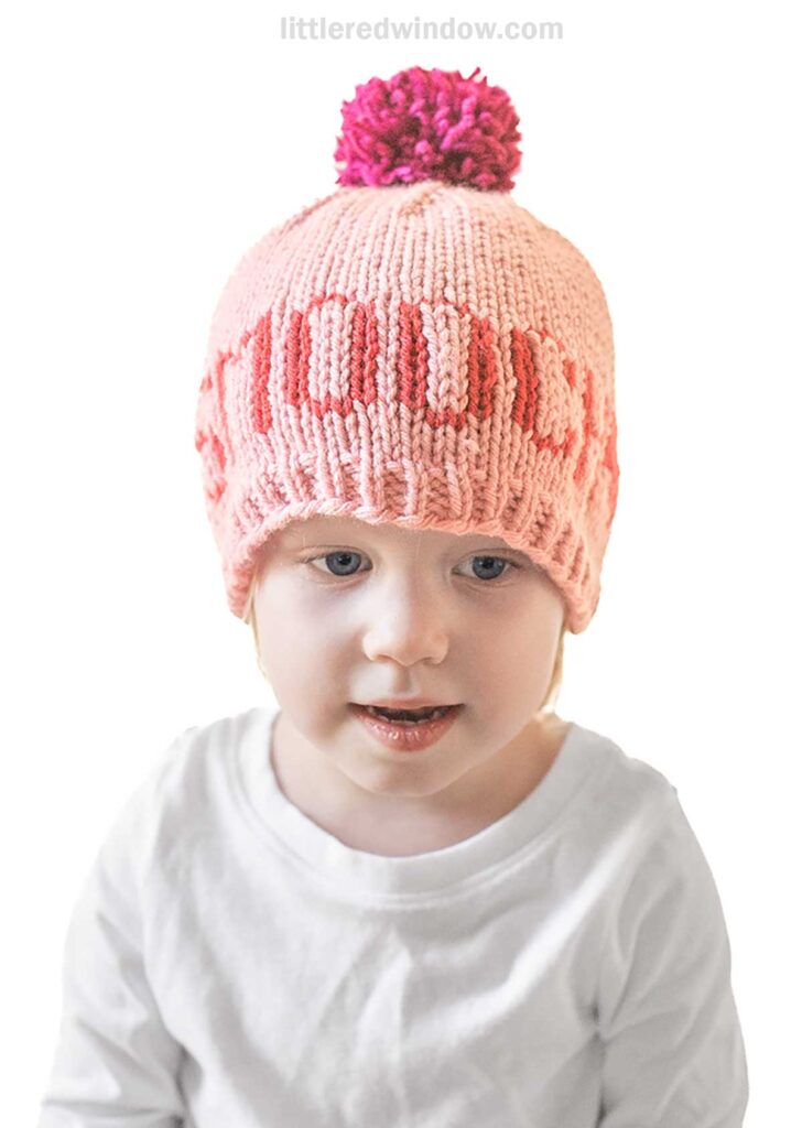 A young child sports a charming pink knit hat from Love Notes Hats, highlighted by a red pom-pom and delightful design around the crown. Dressed in a crisp white shirt, their neutral expression contrasts with the light background, creating an endearing portrait.