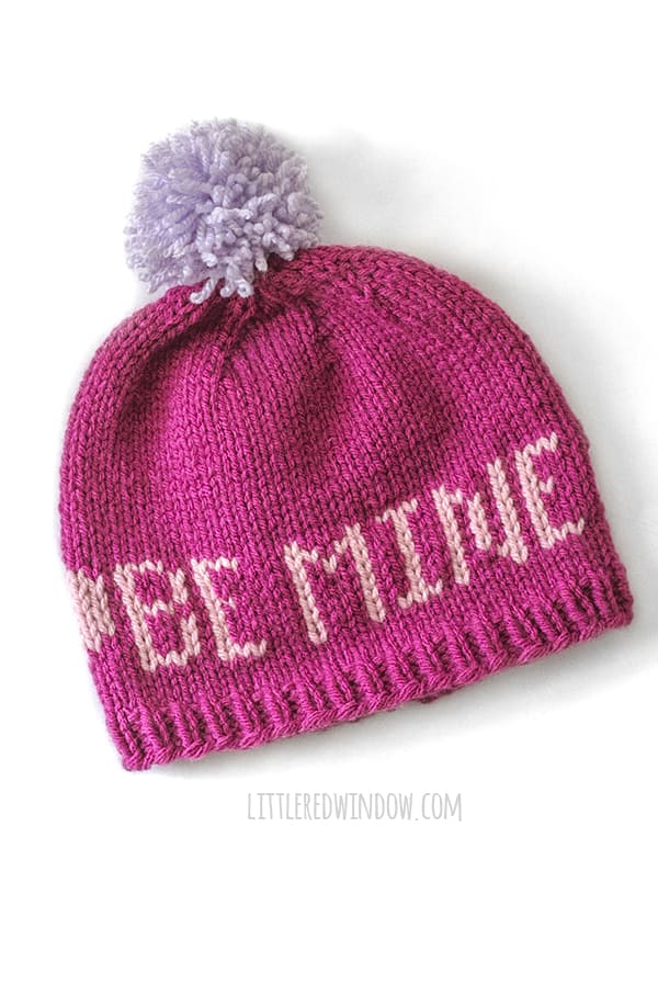 Magenta knit hat with light purple pom pom and light pink words that say BE MINE on a white background