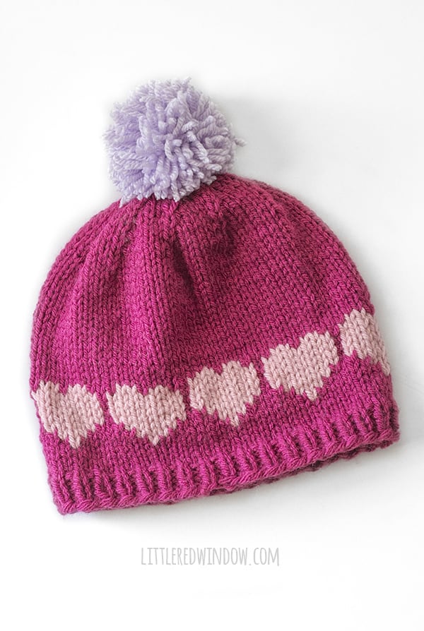 Magenta knit hat with light purple pom pom and light pink heart shapes