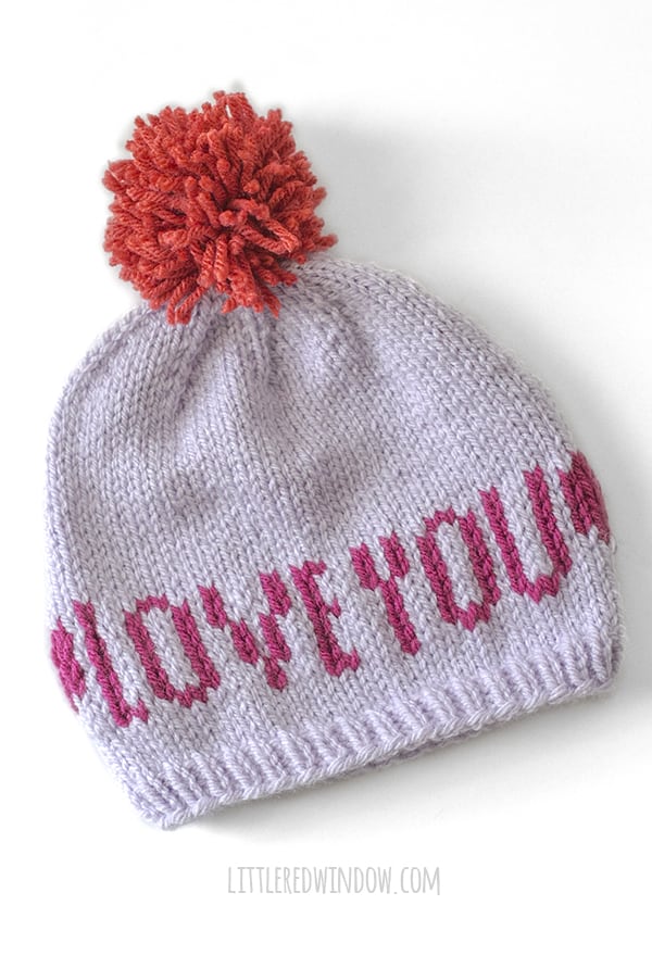 lavender knit hat with red pom pom on top and magenta words that say LOVE YOU flat on a white background