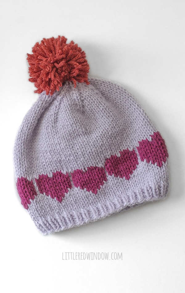 lavender knit hat with red pom pom on top and magenta hearts in a row flat on a white background