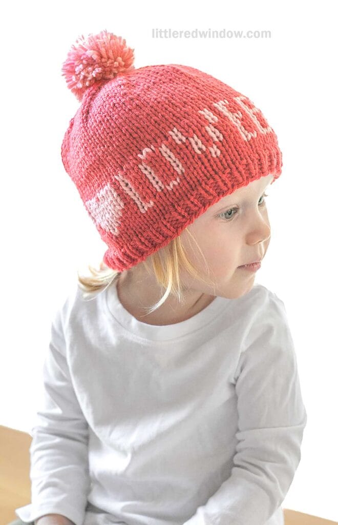 A child wearing a red Love Notes hat, adorned with a pink pom-pom, sits looking to the side. This charming accessory features the words