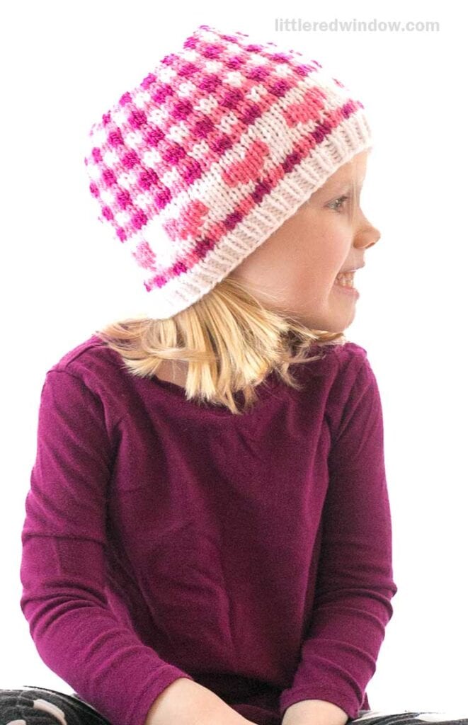 A young child wearing a purple shirt sits smiling, looking to the side. She has a pink and white Valentine plaid knit hat with heart patterns. The background is plain and bright.
