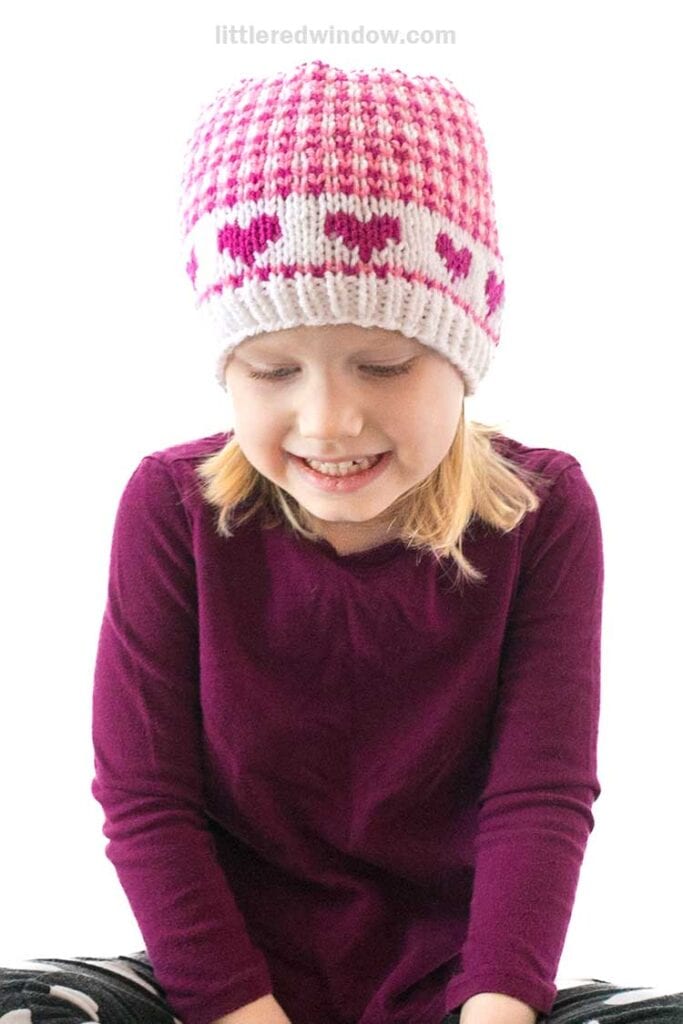 A child wearing a pink and white knitted beanie with heart patterns, reminiscent of Valentine-themed plaids, and a maroon sweater is smiling and looking down. The background is a crisp white.