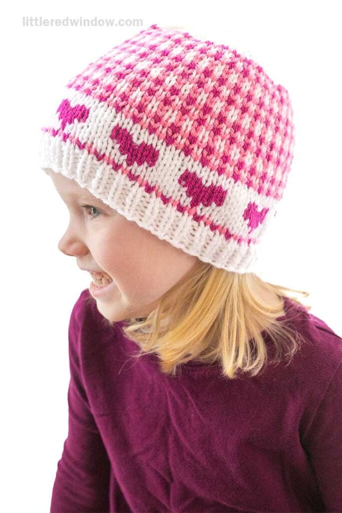 A child wearing a pink and white knitted hat with heart patterns, reminiscent of Valentine charm, is smiling and looking to the side. The child is also wearing a dark purple top.