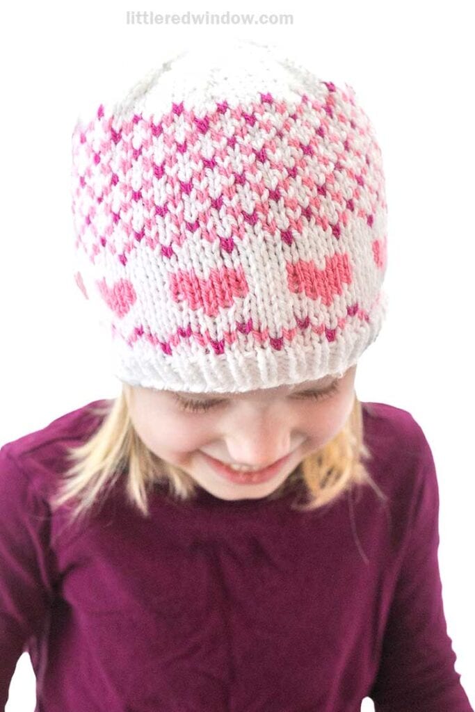 A child in a knitted hat adorned with pink and white heart patterns, reminiscent of Valentine designs, smiles while glancing down, dressed in a deep purple top.
