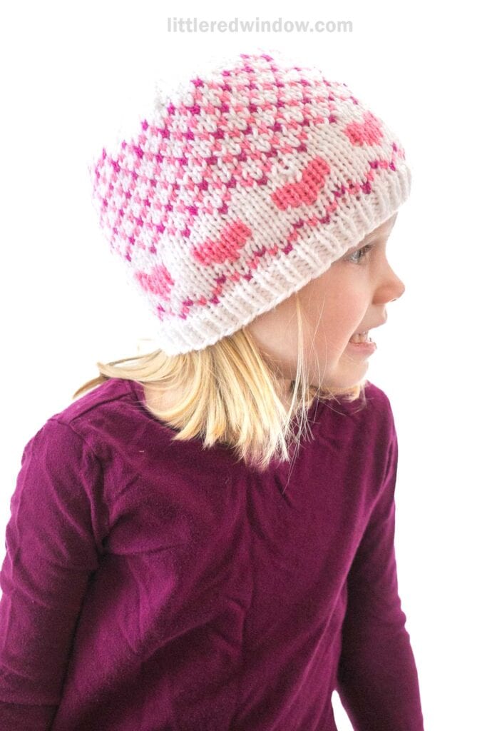 A child wearing a purple shirt and a white knit hat adorned with pink patterns, including bows, smiles warmly while looking to the side. The background is a crisp white, reminiscent of Valentine plaids.