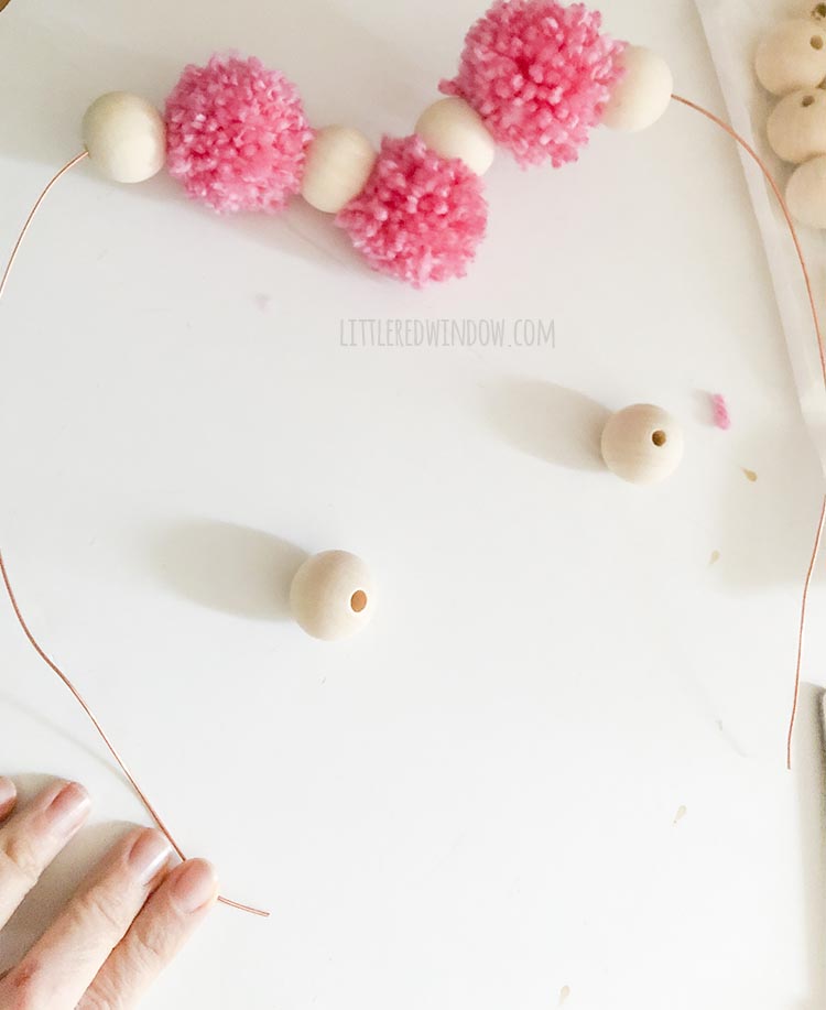 wire heart beginning to be strung with alternating pink yarn poms and wood beads