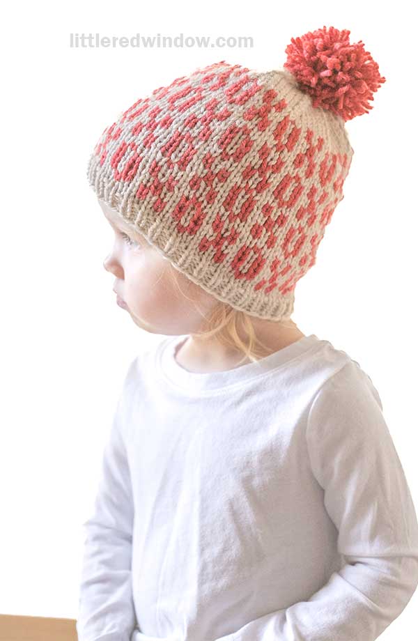 A child wearing a white shirt and a Tic Tac Toe Hat featuring a red and white geometric xoxo pattern with a red pom-pom on top. The child gazes to the side against a light background.