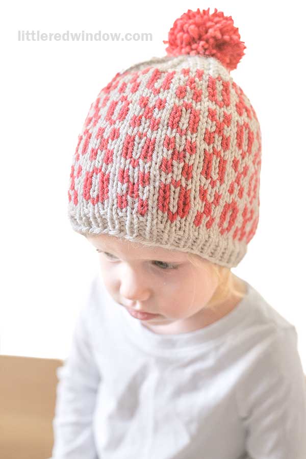 A child wearing a Tic Tac Toe Hat, featuring a whimsical red and gray xoxo pattern with a playful red pom-pom on top. The child, dressed in a light gray shirt, gazes downward thoughtfully.