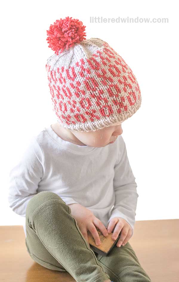 A child wearing a red and white knit Tic Tac Toe Hat with a playful pom-pom sits on a wooden surface. Dressed in a white long-sleeve shirt and olive green pants, the child gazes down to the side while holding a small brown object.