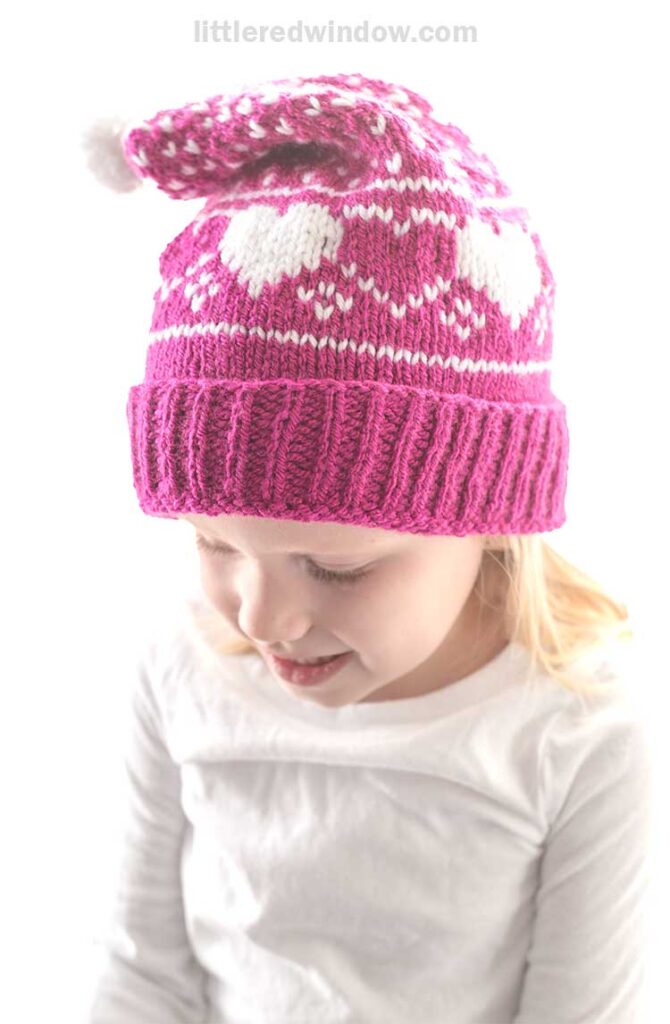 A young child wearing a white shirt and a pink heart stocking cap adorned with heart patterns and a white pompom at the tip. The child is looking down, smiling slightly.