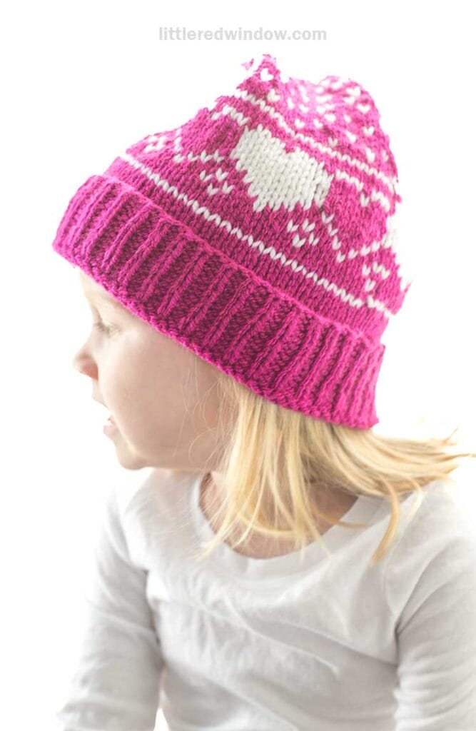 A child wearing a heart stocking cap in bright pink with white hearts and geometric patterns looks to the side. The little one has long blonde hair and is dressed in a white long-sleeve shirt. The background is light and softly blurred, creating a serene atmosphere.