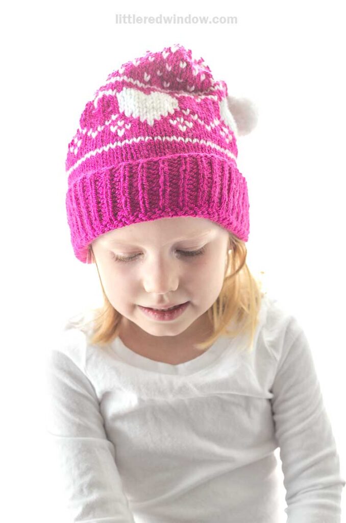 A child wearing a heart-patterned pink stocking cap with white designs and a pom-pom on top. Their light brown hair peeks out as they gaze downward, dressed in a crisp white shirt.