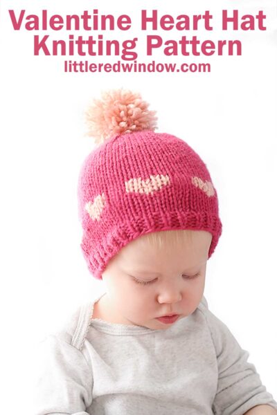 A baby wearing a charming pink heart hat with a pom-pom looks down, wrapped in a light-colored shirt. Above, the text announces, "Valentine Heart Hat Knitting Pattern littleredwindow.com.