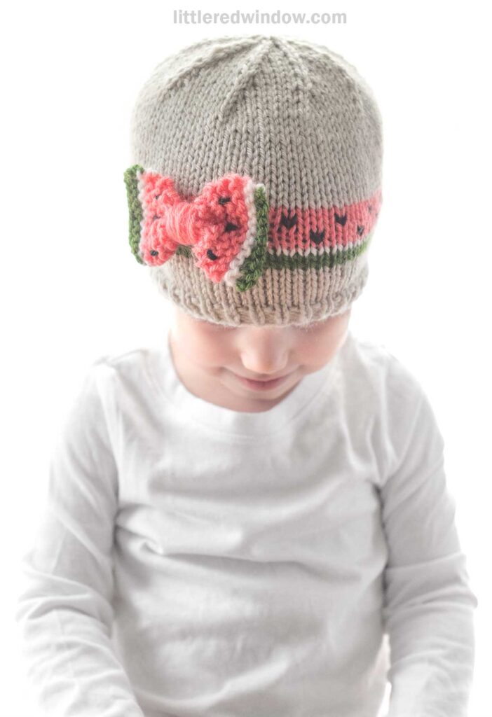 A young child wearing a light gray bow hat with a large pink crocheted bow and green ribbon pattern sits facing downward. The child is dressed in a plain white long-sleeve shirt. The background is plain white, seamlessly highlighting the intricate knitting pattern of the hat.