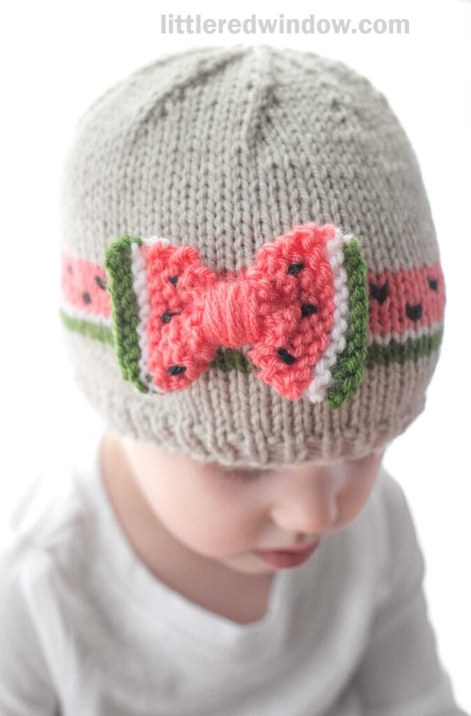 A young child wearing a light gray bow hat with a crocheted pink and green watermelon-themed band. The child's face is partially visible, and they are dressed in a light-colored outfit. The text